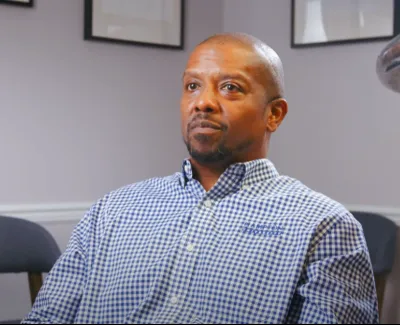 Man in blue and white checkered shirt sitting in a room with framed pictures on the wall, looking attentive.