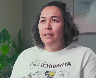 Woman with dark hair wearing a white CoCo Ichibanya t-shirt sitting indoors with green plants in the background