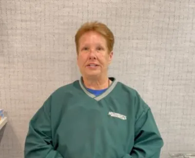 Middle-aged person wearing a green Mariners sweatshirt sitting indoors against a light patterned wall.