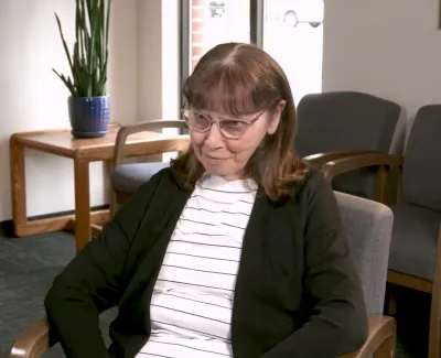 Middle-aged woman with glasses sitting in a gray chair in a room with wooden furniture and plants.
