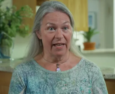 Middle-aged woman with gray hair wearing a patterned blue shirt speaks in a bright kitchen setting.