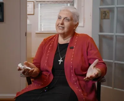 Elderly woman in red cardigan and cross necklace speaking warmly while seated in a cozy room.