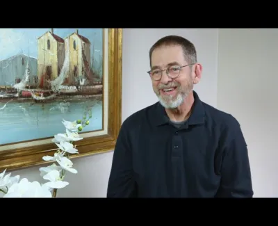 Smiling elderly man with glasses and beard sitting near a framed painting of boats and white orchids.