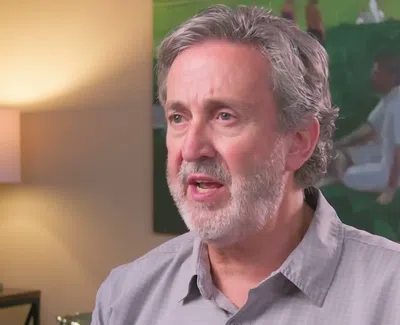 Mature man with gray hair and beard speaking indoors near lamp and painting on the wall