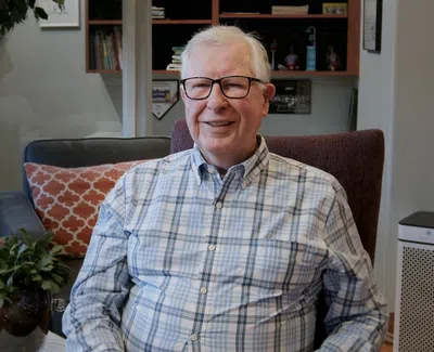 Smiling elderly man in glasses wearing a plaid shirt sitting in a cozy living room with plants and bookshelves.