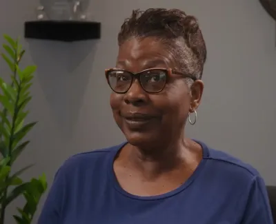 Smiling woman wearing glasses and a blue shirt sitting indoors with plants and decorative items in the background