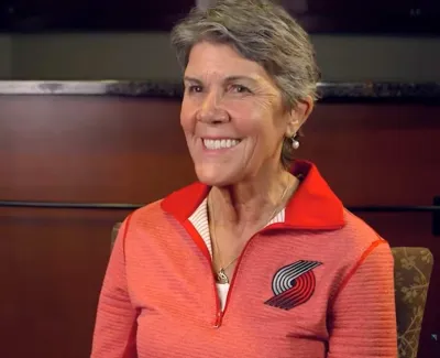 Smiling older woman wearing a Portland Trail Blazers pullover sitting in an indoor setting with a dark wood background.