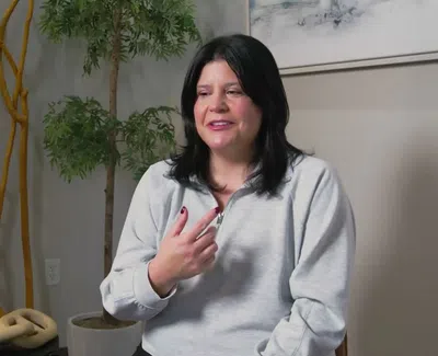 Woman with dark hair in a light gray sweater sitting indoors with plants and decorative chains in the background.