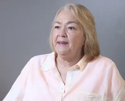 Older woman with blonde hair wearing a light pink shirt sitting against a gray wall and speaking.