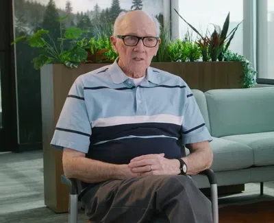 Elderly man wearing glasses and a striped shirt sitting in a modern living room with plants and a sofa behind him.