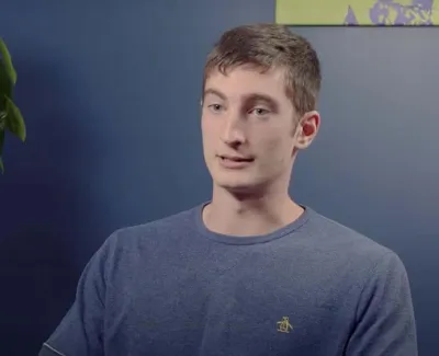 Young man in blue shirt seated indoors next to a braided money tree plant against a two-tone blue wall.