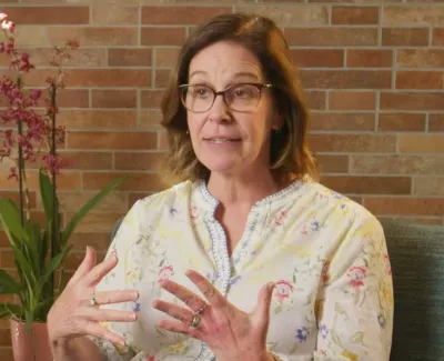 Middle-aged woman in glasses gesturing while speaking with plants and brick wall background