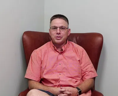 Man in glasses and coral checkered shirt sitting in a brown leather chair against a plain wall.