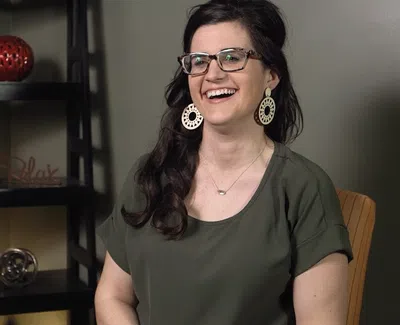 Smiling woman with glasses and large earrings sitting in front of a black shelving unit with decorative items.