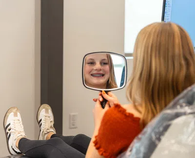 Girl with braces smiling while looking at her reflection in a handheld mirror at orthodontist office.