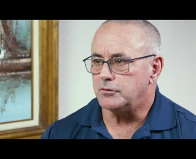 Middle-aged man with glasses wearing a navy blue polo shirt, sitting indoors near framed art on wall.