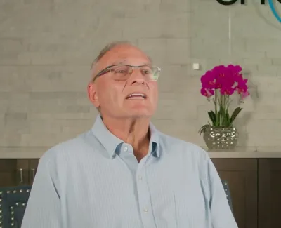 Elderly man with glasses speaking in a modern dental office with orchid and logo background.
