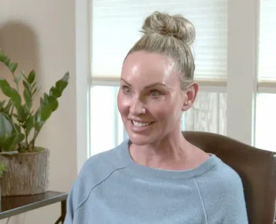 Smiling woman with blonde hair in a bun sitting in a chair near a plant and window blinds in a cozy room.