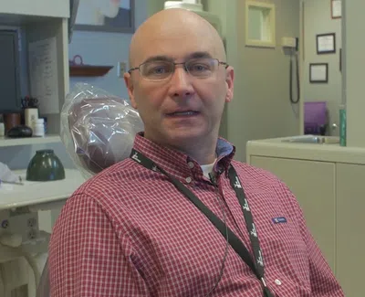 Man wearing glasses and red checkered shirt sitting in a dental office with dental equipment in the background