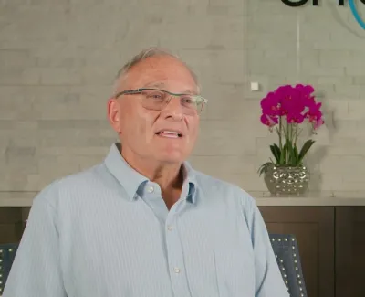 Elderly man with glasses speaking in a modern dental office with pink orchids and logo wall art.