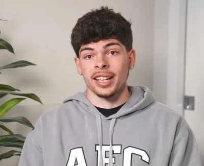 Young man wearing grey AEO hoodie speaking indoors next to a large green potted plant.