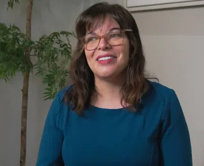 Smiling woman wearing glasses and teal dress posing indoors with green plants and wooden decor in background