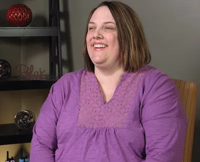 Smiling woman in a purple blouse sitting indoors near a shelf with decorative items and a 'Relax' sign.