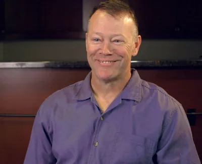 Smiling man wearing a purple shirt sitting indoors with a relaxed posture and warm lighting.