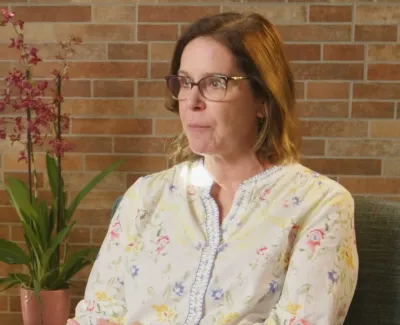 Middle-aged woman with glasses sitting indoors near green plants and a brick wall background.