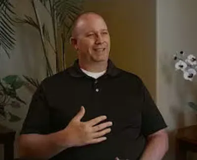 Smiling man in black polo shirt seated indoors with plants and a table holding an orchid and award plaque.