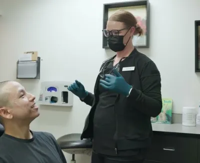 Dental professional wearing mask and gloves talking to smiling male patient in clinic room