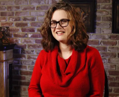 Smiling woman with glasses wearing a red sweater sitting indoors against a brick wall with wooden furniture.