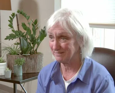 Smiling elderly woman with white hair wearing a blue shirt sitting in a cozy room with plants and natural light.