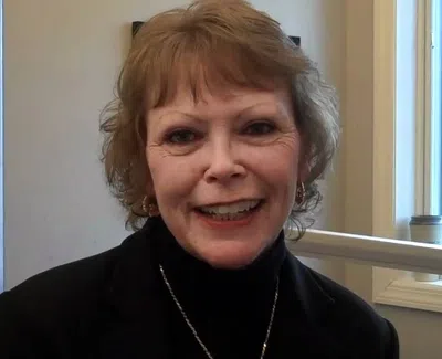 Smiling woman with short hair wearing a black top and necklace in a bright room with large windows.