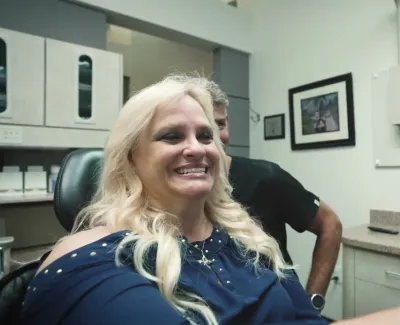 Smiling woman with blonde hair sitting in a dental chair taking a selfie with a man in a dental office.