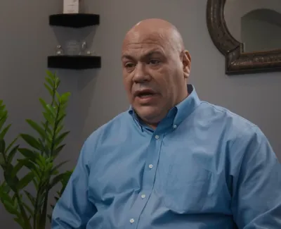 Bald man in blue shirt speaking in a modern office with plants and mirror in the background.