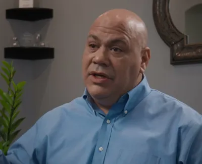 Bald man in blue shirt speaking and gesturing with hand in a modern office setting with plants and awards.