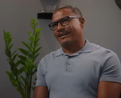 Man wearing glasses and a light blue polo shirt smiling indoors with plants and office decor in the background
