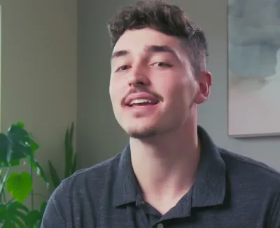 Young man with short hair and mustache smiling indoors near green plants and window light.