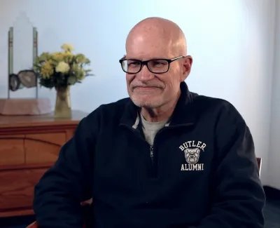 Smiling bald man with glasses wearing a Butler Alumni sweatshirt sitting in a room with wooden furniture and plants.