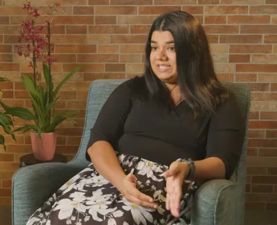 Woman in black top and floral skirt sitting in blue chair gesturing while talking against brick wall