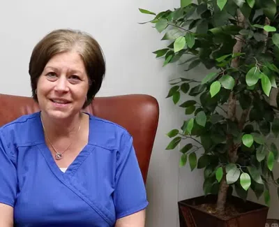 Smiling woman in blue scrubs sitting in brown chair next to a potted green plant against a white wall.