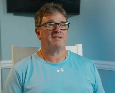 Man wearing glasses and blue Under Armour shirt sitting indoors against light blue wall during interview