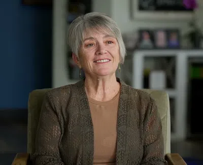 Smiling older woman with short gray hair wearing a brown top and cardigan sitting in a modern room.