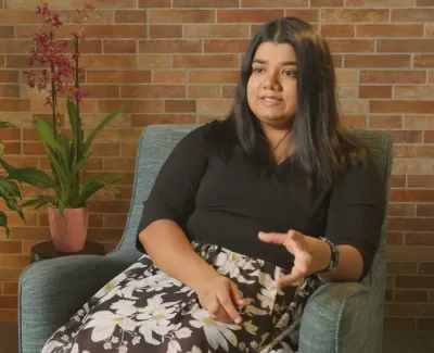 Woman in floral skirt and black top sitting in blue chair, speaking against a brick wall backdrop with potted plants