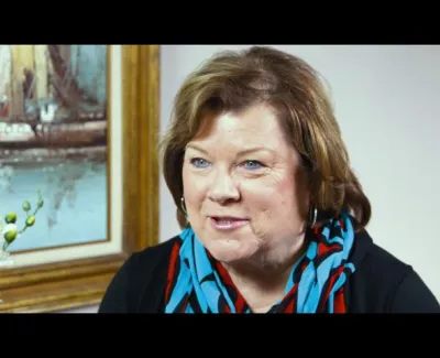 Middle-aged woman with short brown hair wearing a colorful scarf, smiling indoors next to a framed painting.