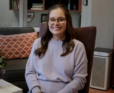 Smiling woman with glasses in a cozy living room sitting on a chair near a plant and patterned pillow.