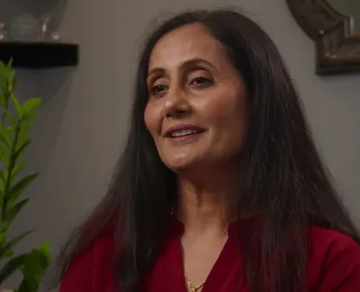 Smiling middle-aged woman with long hair wearing a red top sitting indoors with green plant in background.