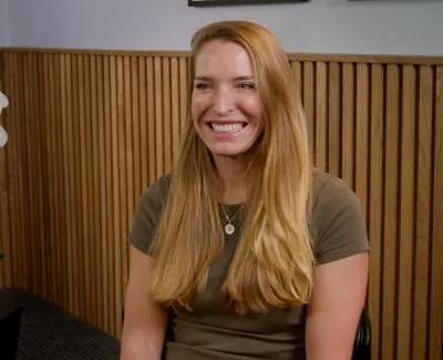 Smiling woman with long blonde hair sitting near a white orchid plant in a modern wooden-paneled room.