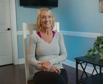 Smiling woman seated in a cozy room with light blue walls and a table holding a plant and a decorative smile sign.